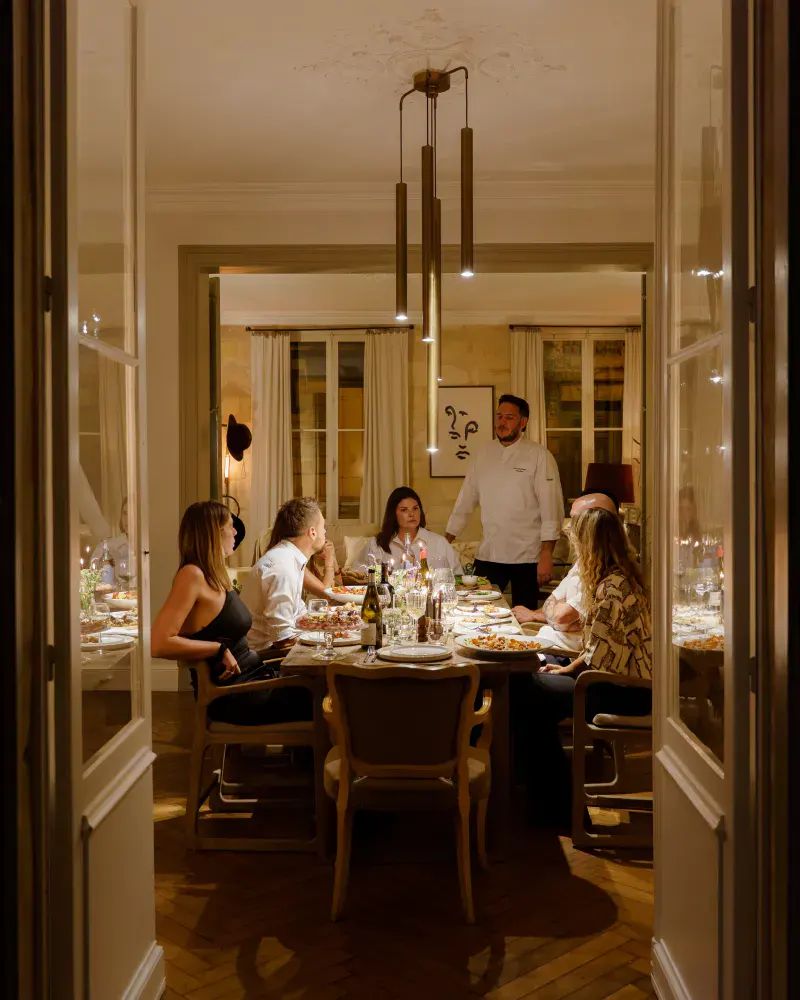 Diner avec plusieurs personnes autour d'une table et un chef debout à côté. L'ambiance est tamisée et intimiste.